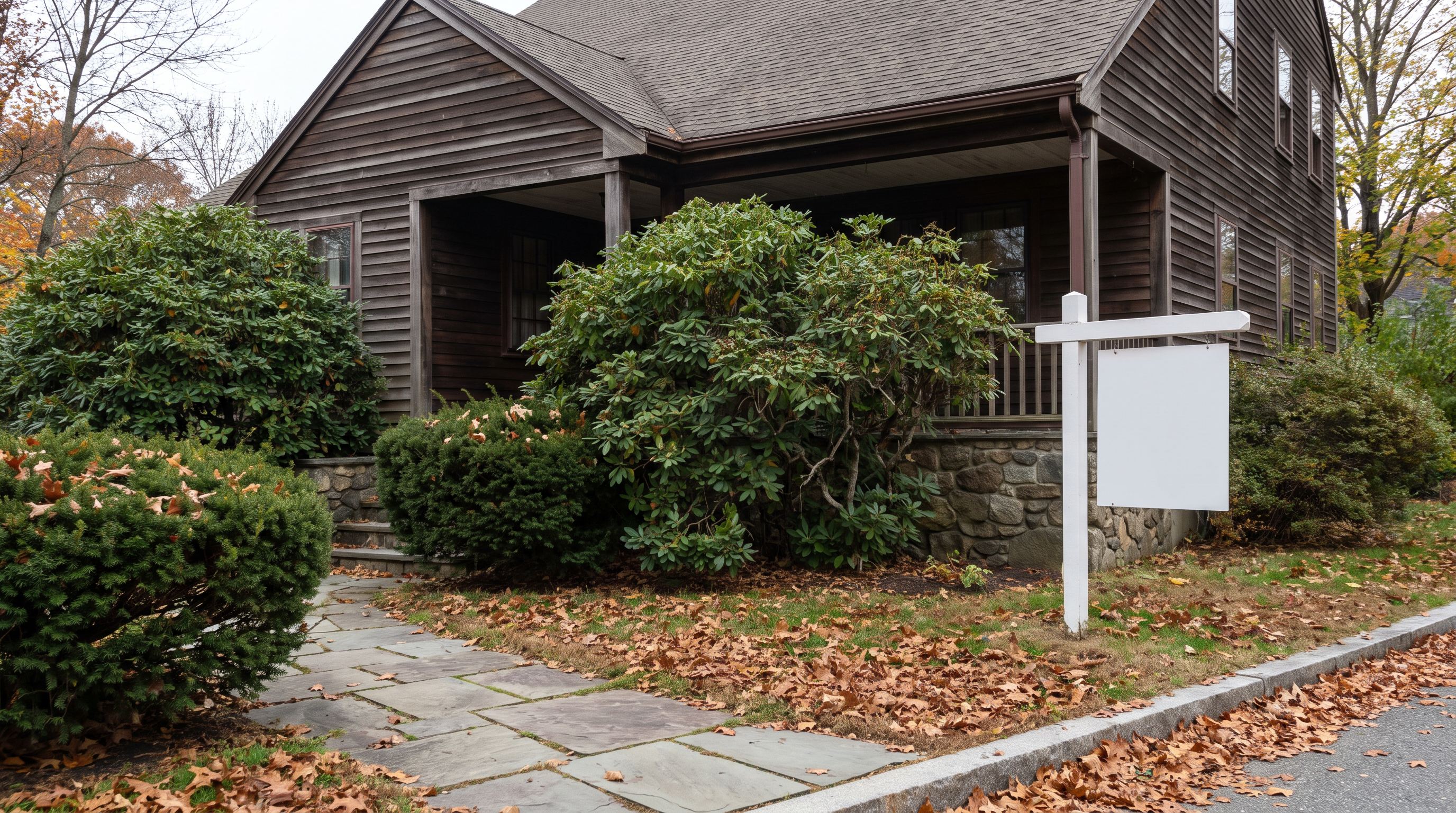 An empty older New England house seen from the front walk on an overcast autumn morning, overgrown shrubs and a for sale sign at the curb, no people in frame